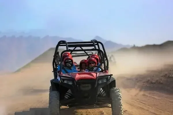 A red dune buggy with two helmeted passengers drives toward the camera along a dusty desert track with mountains in the background.