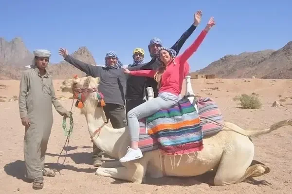 Two tourists ride camels led by a Bedouin guide across the desert with mountains in the background; a website watermark is visible at the bottom of the photo.