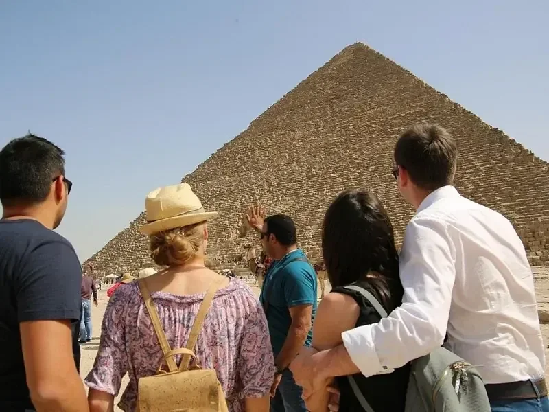 A small group of tourists, seen from behind, look toward the Great Pyramid of Khufu at Giza under a clear sky.
