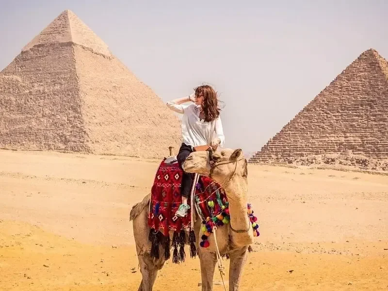 A woman rides a camel between two of the Giza pyramids, with desert sand in the foreground.