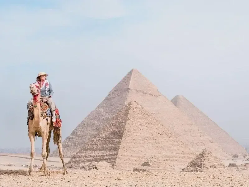 A tourist sits on a camel photographed from the side near the Great Pyramid in hazy desert light.