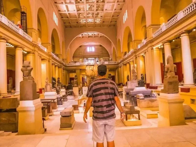 A man walks through the Grand Hall of the Egyptian Museum, surrounded by large granite statues and stone sarcophagi.
