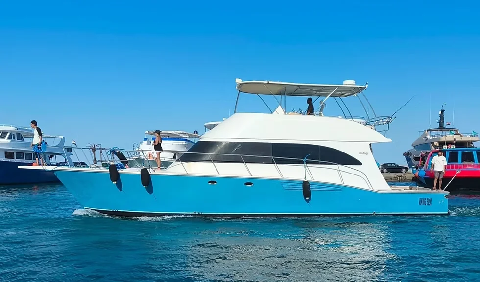 A turquoise-hulled motor yacht named 'Viking Sun' cruises near a marina with other boats moored nearby.