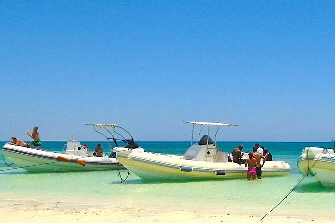 Several small motorboats are anchored in shallow turquoise water near a white sand beach, with tourists wading nearby.