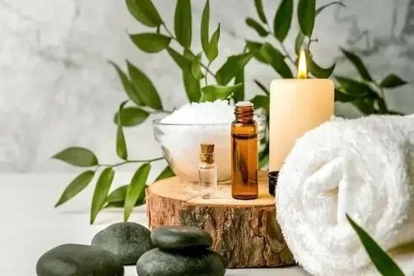 A spa still-life of bath salts in a glass bowl, small amber oil bottles, a lit candle, black hot stones, and a rolled white towel arranged on a wood slab with greenery.