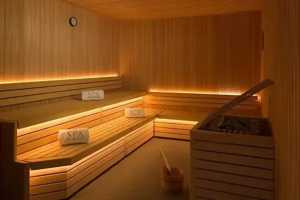 A wood-paneled sauna room with illuminated tiered benches holding folded towels labeled SPA, and a stove with a wooden bucket in the corner.