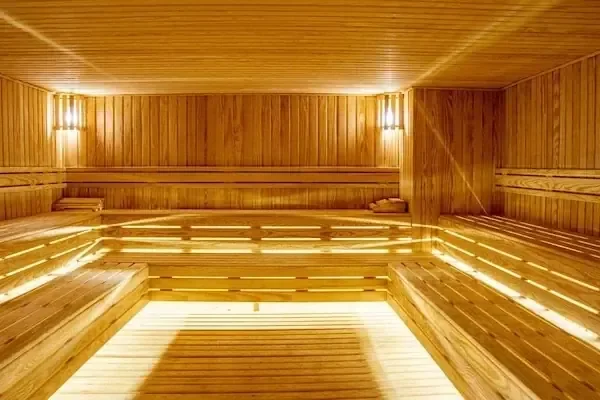 A wide-angle view of an empty wooden sauna with tiered benches along three walls and wall-mounted lamps casting a warm glow.