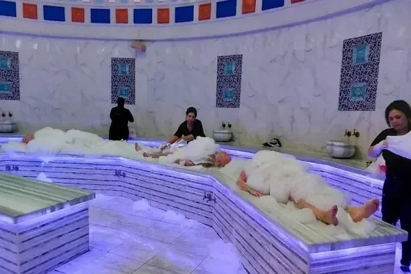 Several people wrapped in white foam and towels lie on a heated marble hammam platform in a domed tiled room, attended by two staff members.
