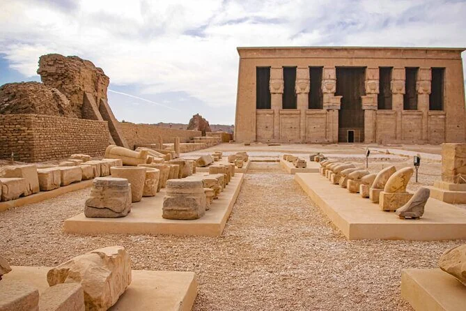 Scattered stone column fragments and ruins lie in the courtyard in front of Dendera Temple's facade.