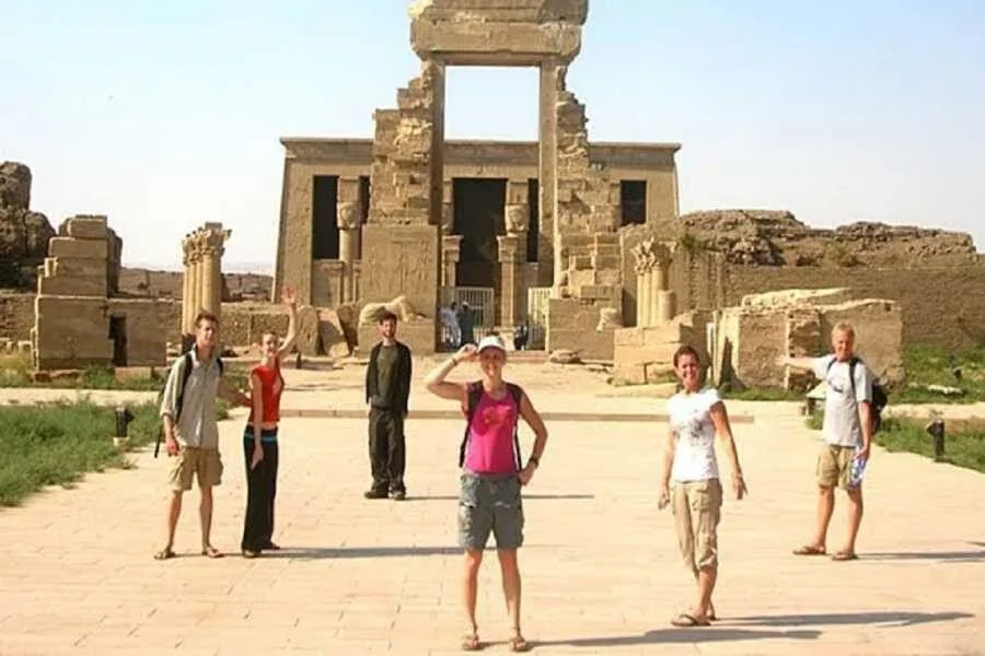 Six tourists pose and wave in front of the ruined stone gateway of a temple entrance.