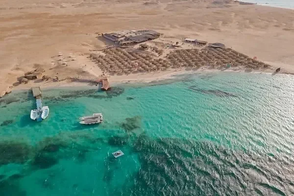Aerial view of a private beach island with straw umbrellas, a jetty, and boats anchored in turquoise water.