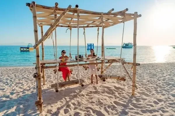 Couple sitting at a bamboo beach swing-table set on white sand by the sea at sunset.
