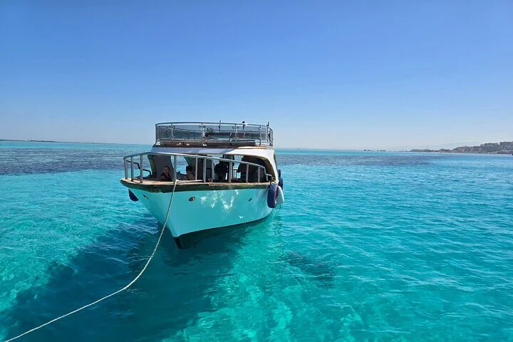 White glass-bottom boat anchored in shallow turquoise water with an empty upper deck and coastline in the distance.