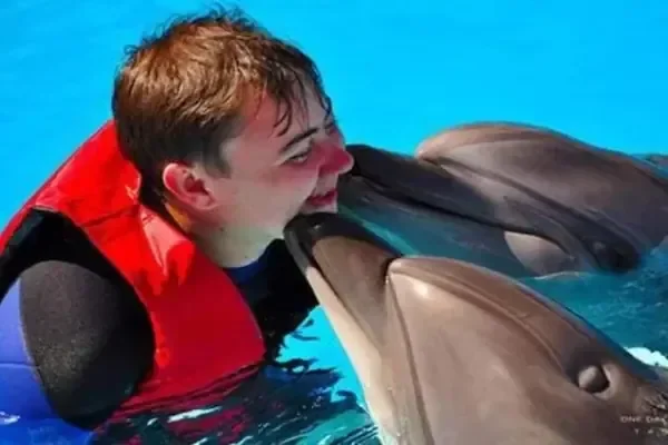 Boy in a red life vest smiling as a dolphin nuzzles his cheek in a pool.