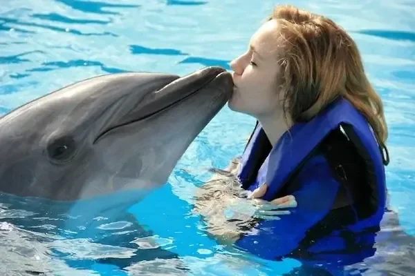 Woman in a blue life vest kissing a dolphin on the mouth in a pool.