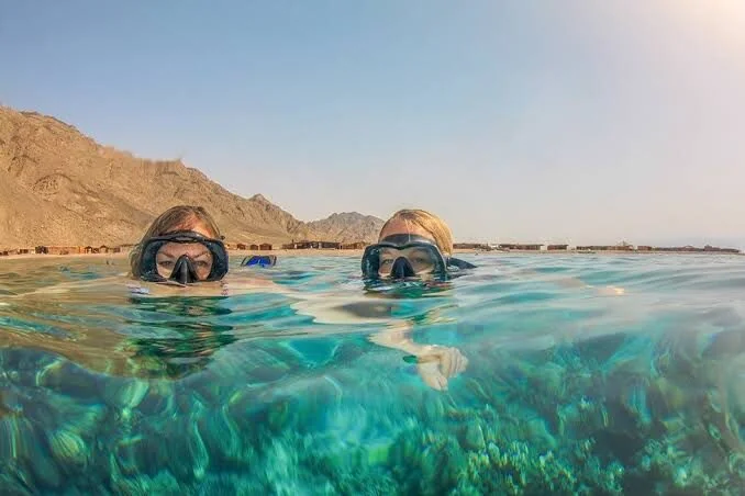 Two snorkelers' faces at the water's surface with a rocky mountain coastline behind them and coral visible underwater.