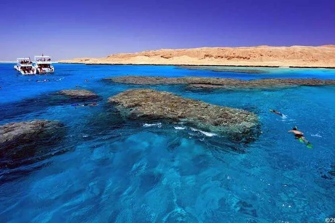 Aerial view of coral reef patches in bright blue water near a sandy island shoreline, with a boat and swimmers visible.