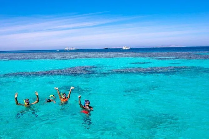 Three snorkelers with raised arms floating in turquoise water above a reef, boats visible in the distance.