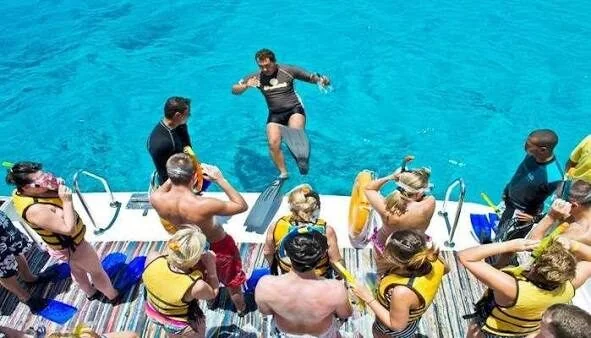 A guide leaps into turquoise water from a boat while guests in life jackets watch and prepare to snorkel.