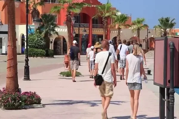Tourists walking along a palm-lined pedestrian promenade past reddish buildings with shops and cafes.