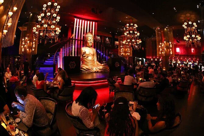 A dimly lit lounge with a large golden Buddha statue, red lighting, chandeliers, and guests seated at tables.