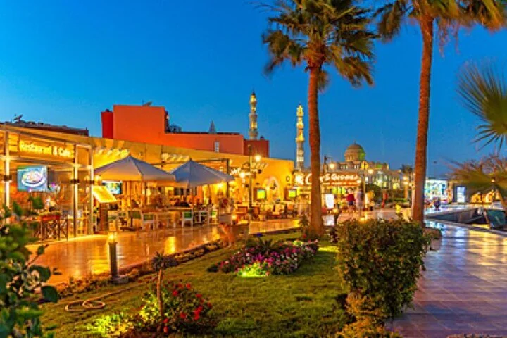An outdoor restaurant terrace with umbrellas and palm trees lit up at dusk, with illuminated minarets visible in the background.