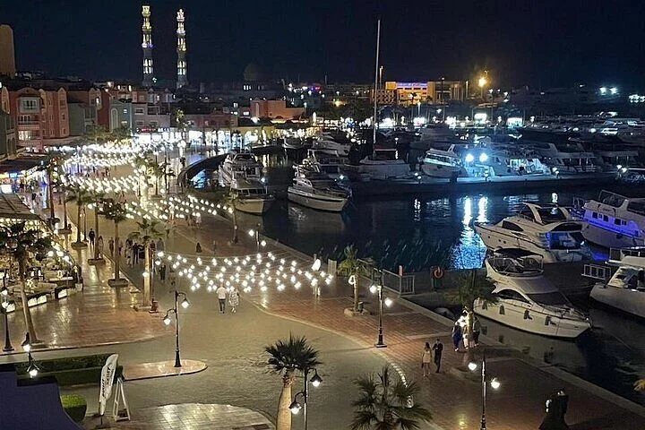 An aerial night view of a marina promenade strung with lights, boats docked along the water, and illuminated minarets beyond.