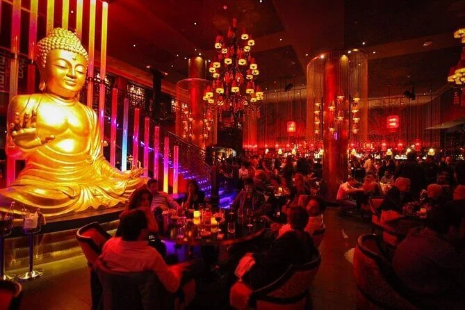 A crowded lounge bar at night with a large golden Buddha statue, red chandeliers, and guests seated around tables.