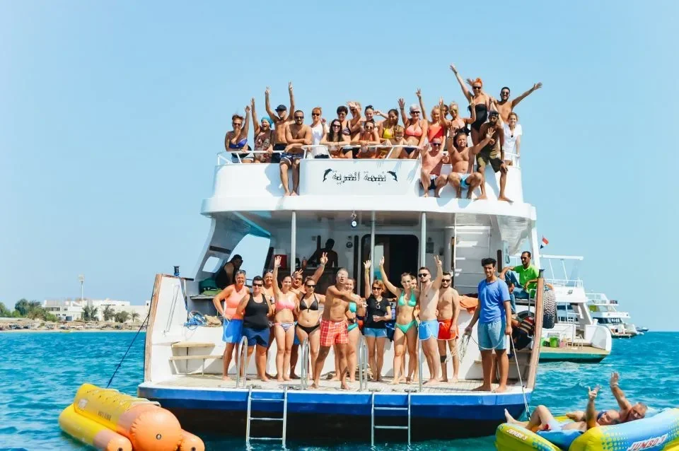 A large tour boat packed with cheering passengers on its upper deck, with more guests posing on the swim platform beside inflatable toys.
