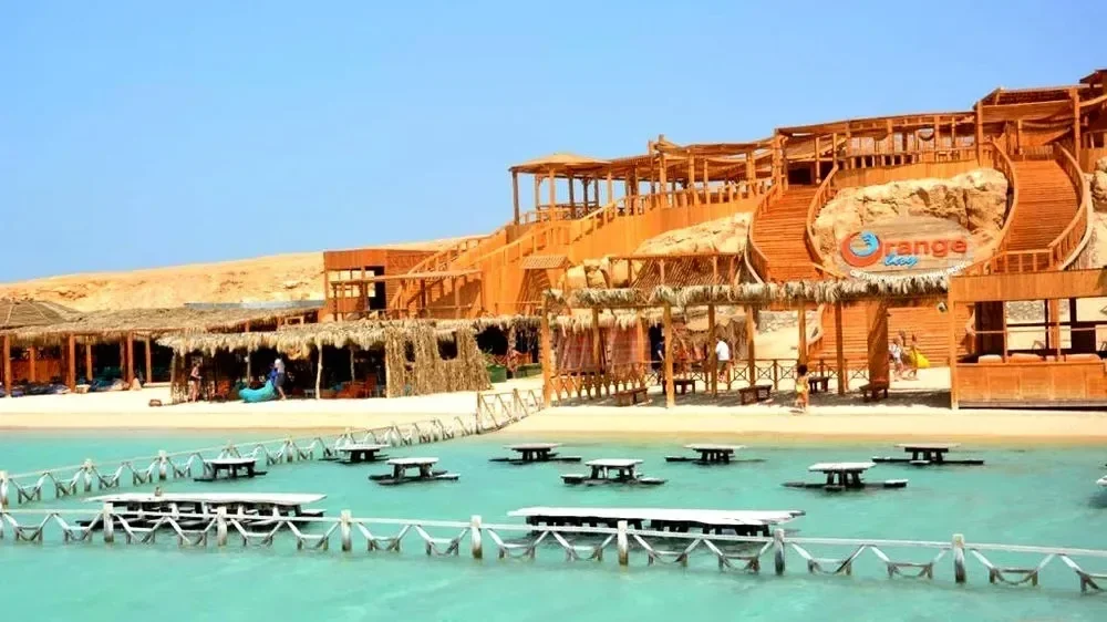 The wooden, multi-level Orange Bay resort building on a sandy hillside with its logo sign, and submerged picnic tables in the foreground sea.