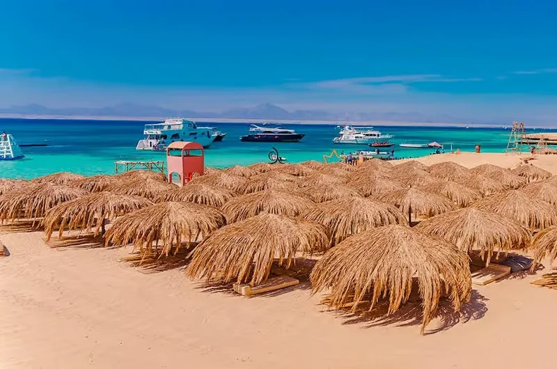 Straw beach umbrellas on golden sand with several tour boats anchored in the turquoise bay and hazy mountains beyond.