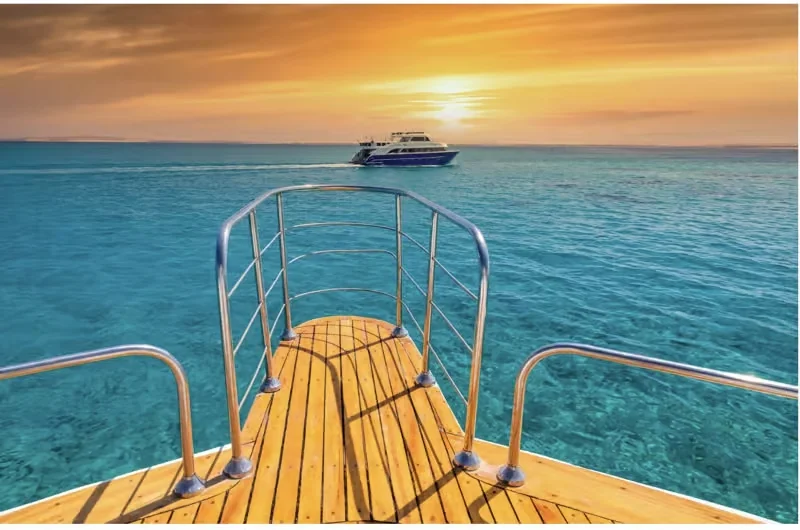 View along a yacht's wooden bow deck toward a second yacht silhouetted against an orange sunset over the sea.