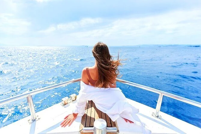 A woman in white relaxes on a yacht's bow seat, her hair blowing in the wind as she looks out over deep blue water.