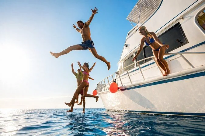 Four friends leap joyfully off the side of a yacht into the sea, caught mid-air against a bright sky.