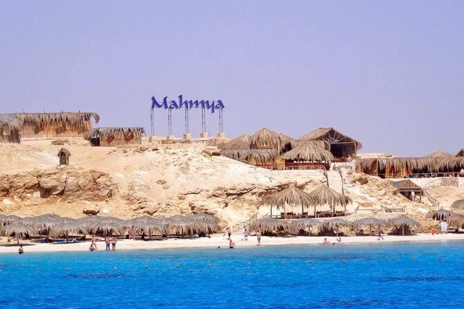 The Mahmya sign rises above sandy cliffs and thatched beach huts, seen from the turquoise water with beachgoers below.