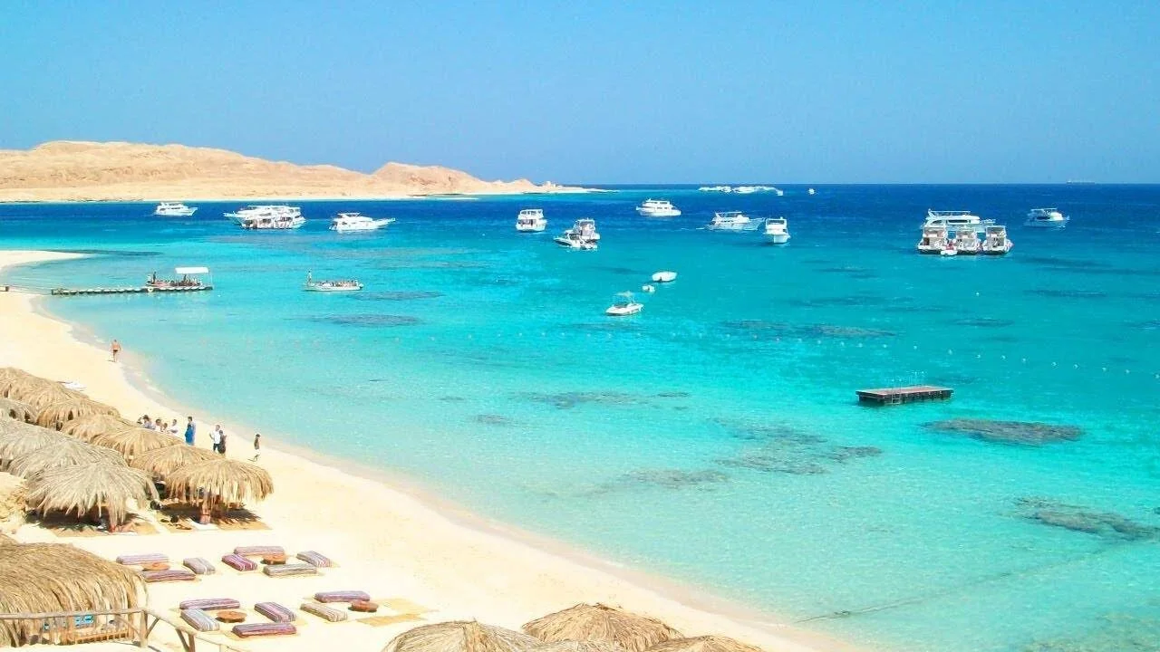 A woman looks out over Mahmya's straw-roofed beach cabanas and crowded white sand toward anchored yachts and a distant island.