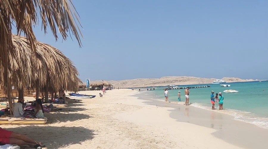 Elevated view of Mahmya's turquoise bay dotted with anchored tour boats, a sandy beach lined with striped loungers under straw umbrellas.