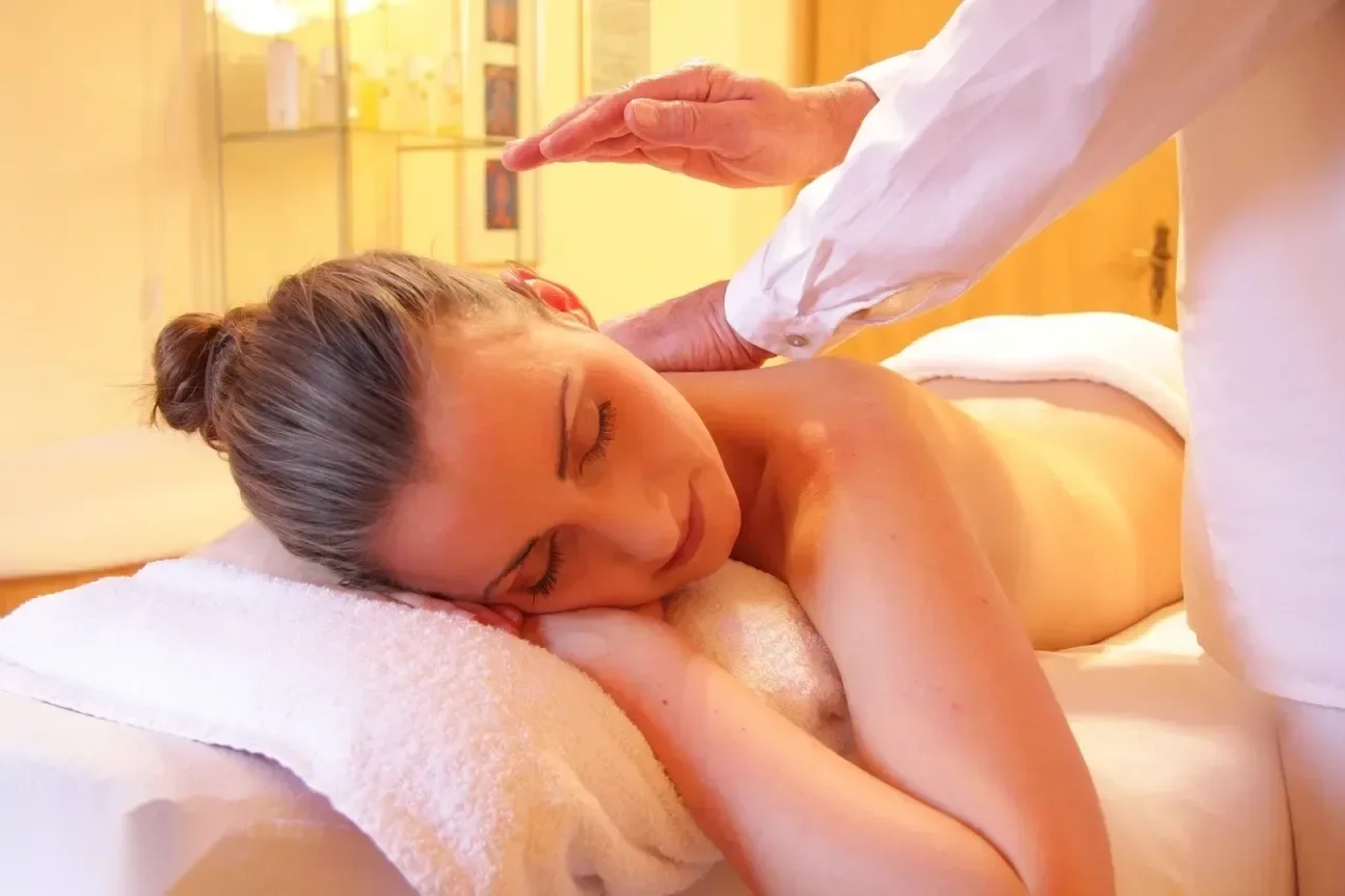 A therapist's hand hovers above a woman's bare shoulder as she lies face-down on a spa treatment table.
