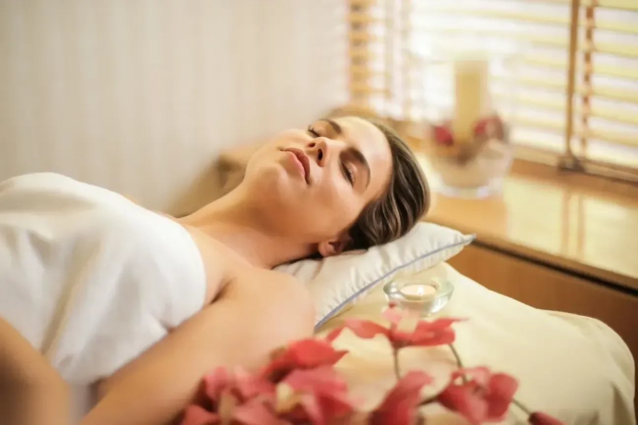 A woman lies back with eyes closed on a spa bed wrapped in a white towel, with red flowers and a lit candle beside her.