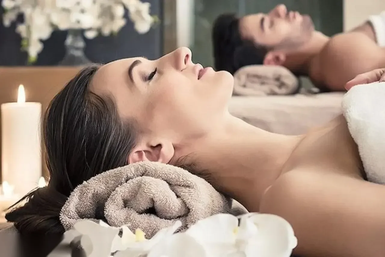 A woman lies back with eyes closed on a spa bed decorated with candles and orchids, with a second guest reflected in a mirror behind her.