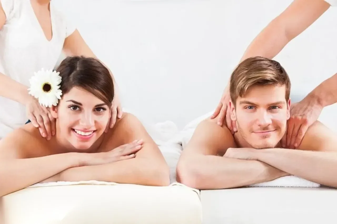 A smiling man and woman lie side by side receiving neck and shoulder massages, with a white flower tucked behind the woman's ear.