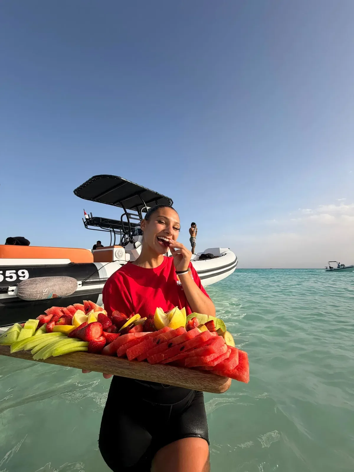 A crew member in a red shirt stands in shallow water holding a large wooden platter piled with watermelon, strawberries, melon, and other fresh fruit, with a speedboat behind her.