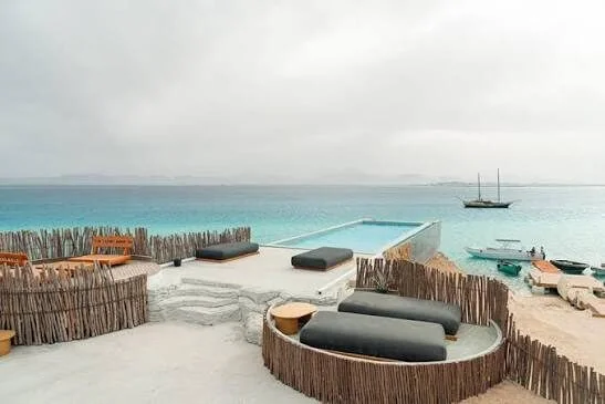 Infinity pool overlooking the turquoise sea with dark cushioned day beds, a sailboat and speedboats moored offshore under an overcast sky.