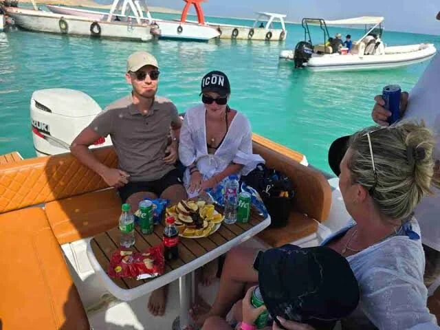 Group of guests seated on a speedboat's cushioned seats eating crisps and orange slices with drinks, other speedboats anchored behind them.