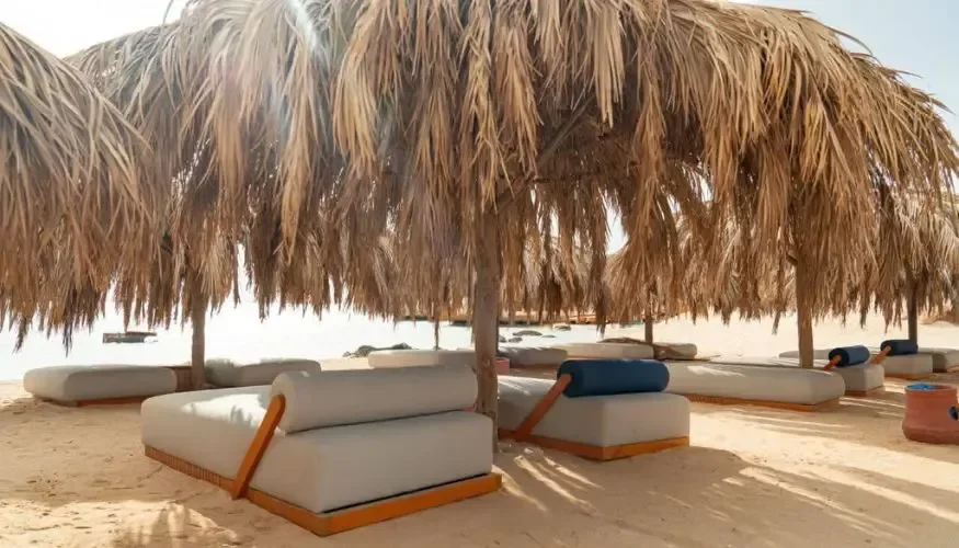 Palm-thatched shade canopy over white and blue cushioned day beds set on the sand facing the water.