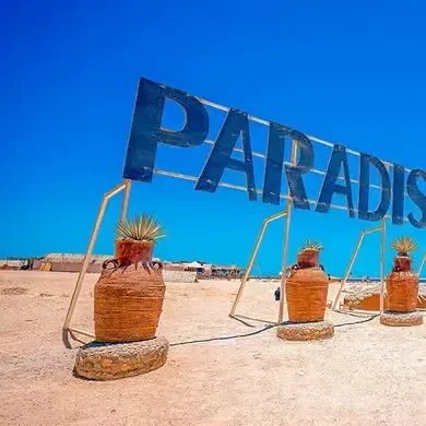 A wooden PARADISE sign stands among terracotta pots on the sand under a clear sky.
