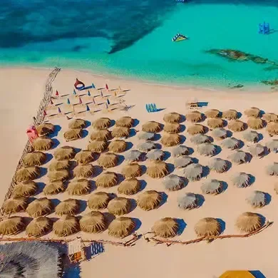 An aerial view shows rows of straw beach umbrellas lined up on white sand beside a turquoise cove.