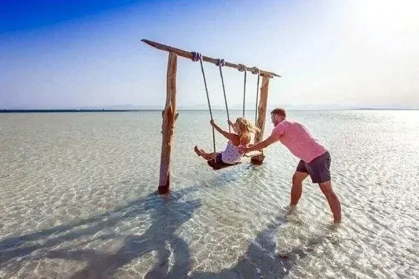 A father pushes a young girl on a wooden swing standing in shallow, crystal-clear turquoise water.