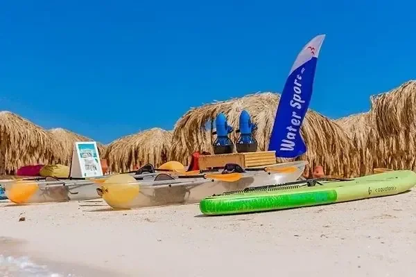 Kayaks, paddleboards and a Water Sport flag are lined up on white sand in front of straw-roofed beach cabanas.