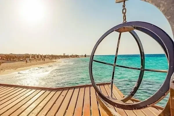 A metal ring frames a view along a wooden pier toward a turquoise beach lined with straw umbrellas.